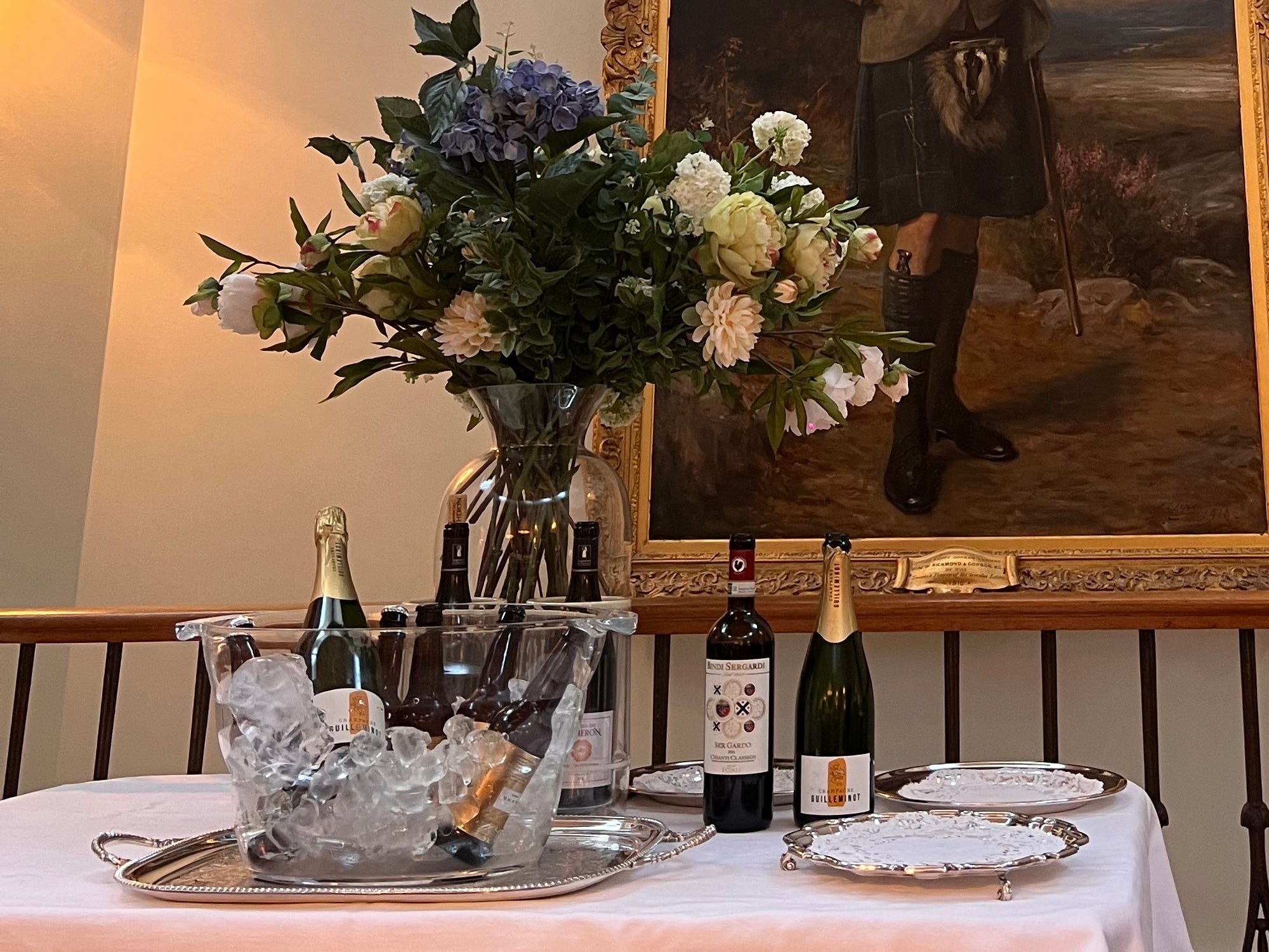 A table topped with bottles of wine, an ice bucket, a vase of flowers, and a painting hanging on the wall in the background.