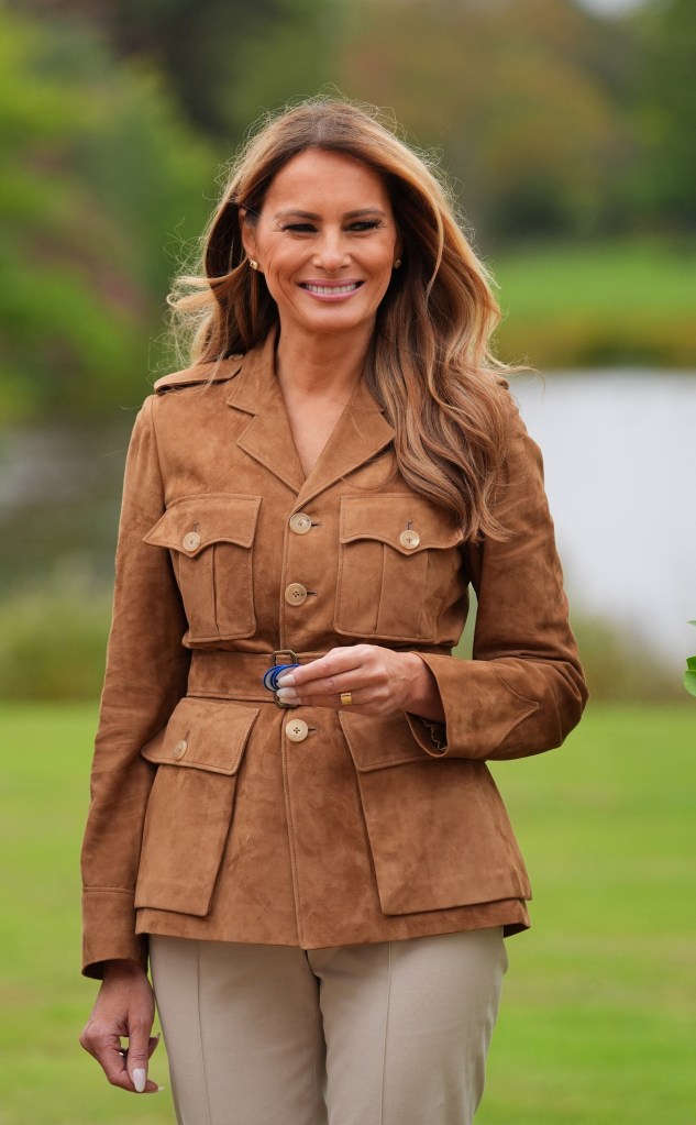 First Lady Melania Trump wearing a brown suede jacket and beige pants, holding a blue badge.