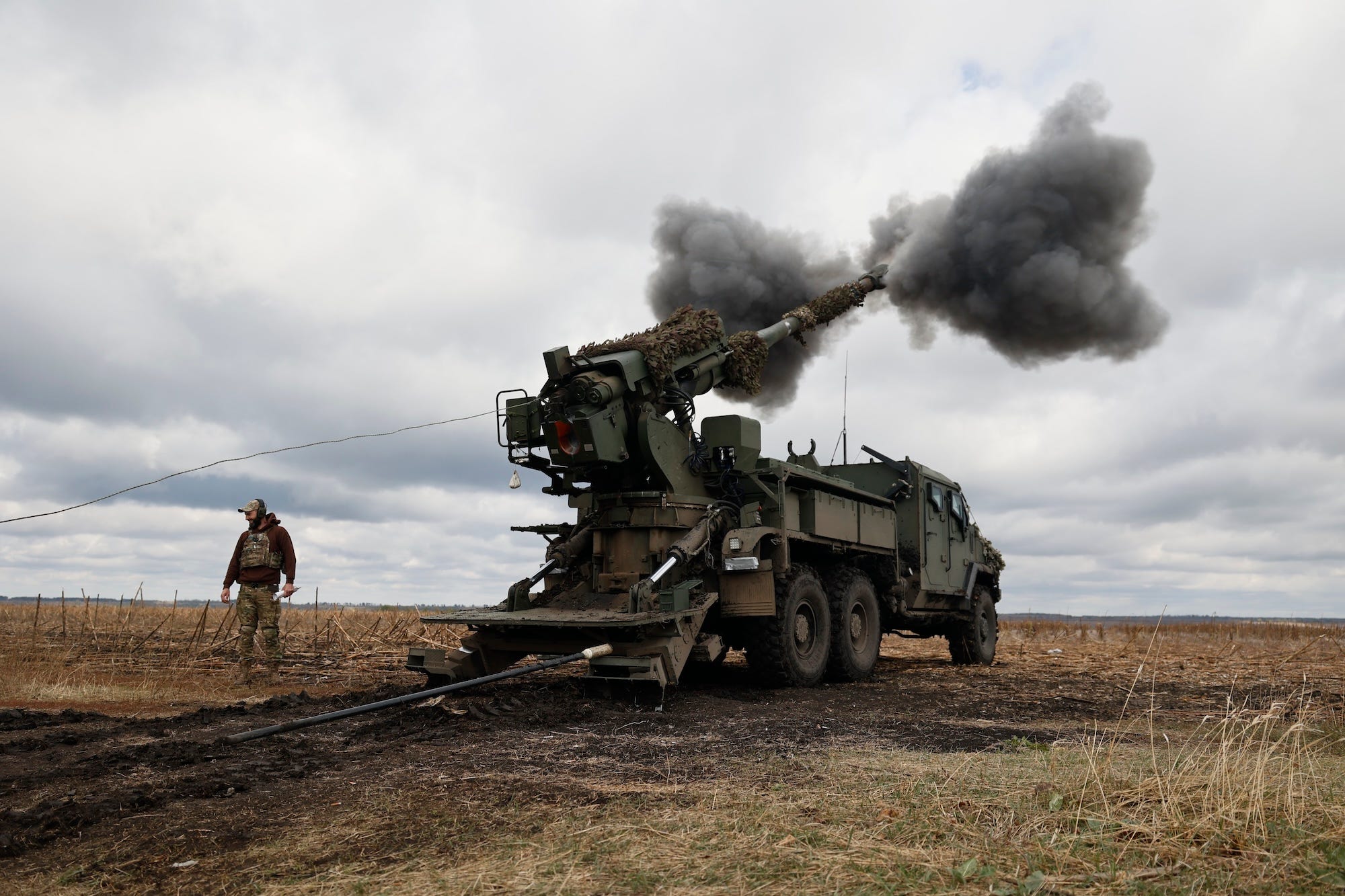 A man in camouflage gear and a black hoodie stands beside a large, green-coloured truck-mounted weapon that has fired and has black smoke around it