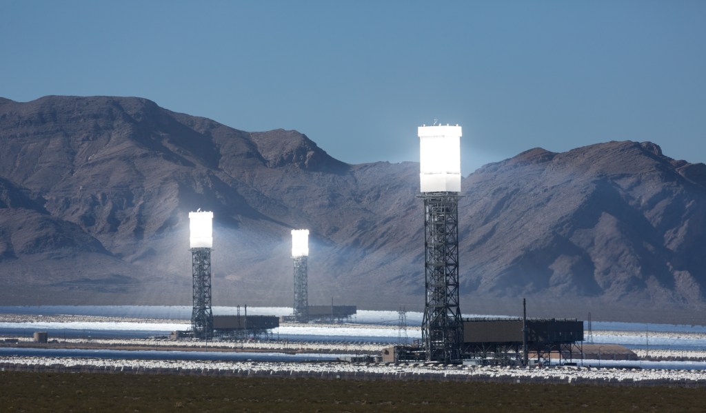 Three large solar thermal towers surrounded by fields of heliostat mirrors with mountains in the background at the Ivanpah Solar Power Facility.