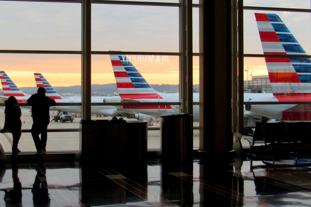 Silhouetted figures looking out a large airport window at American Airlines planes at sunset.