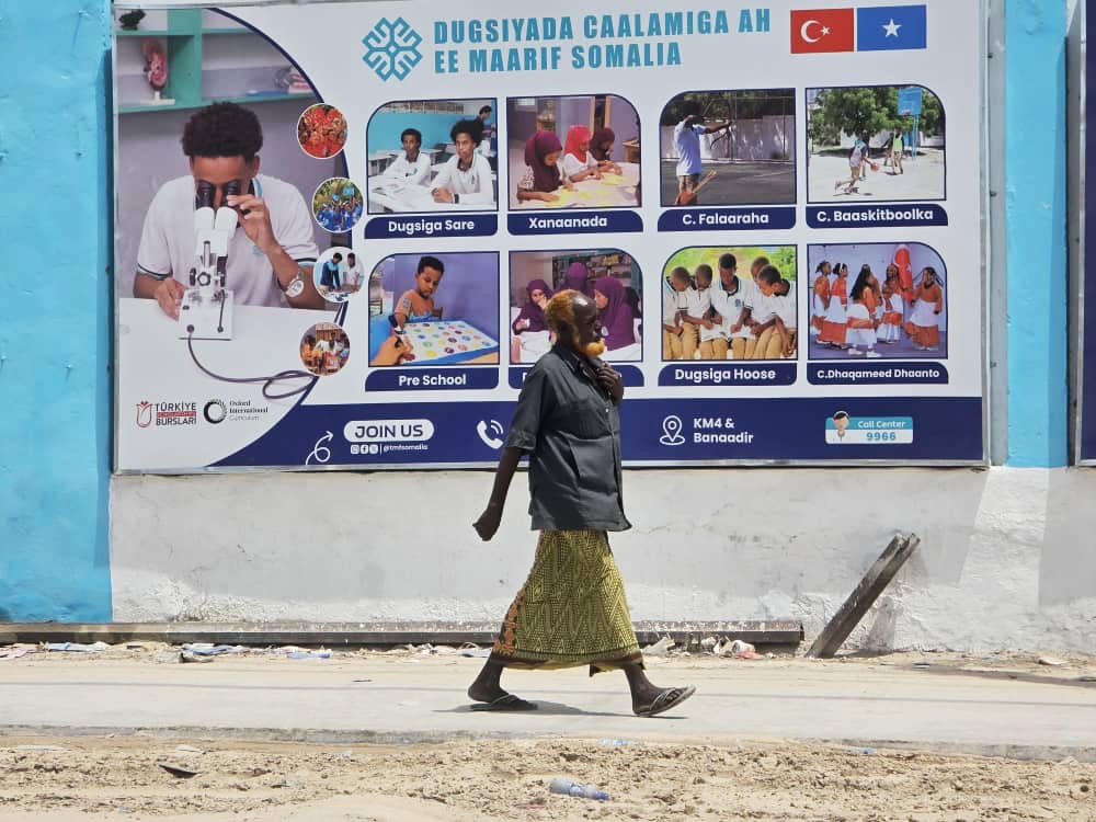 A woman in a headscarf, skirt and long top strides along a path past a wall with a large sign showing multiple photos of students along with Somali text on a wall.