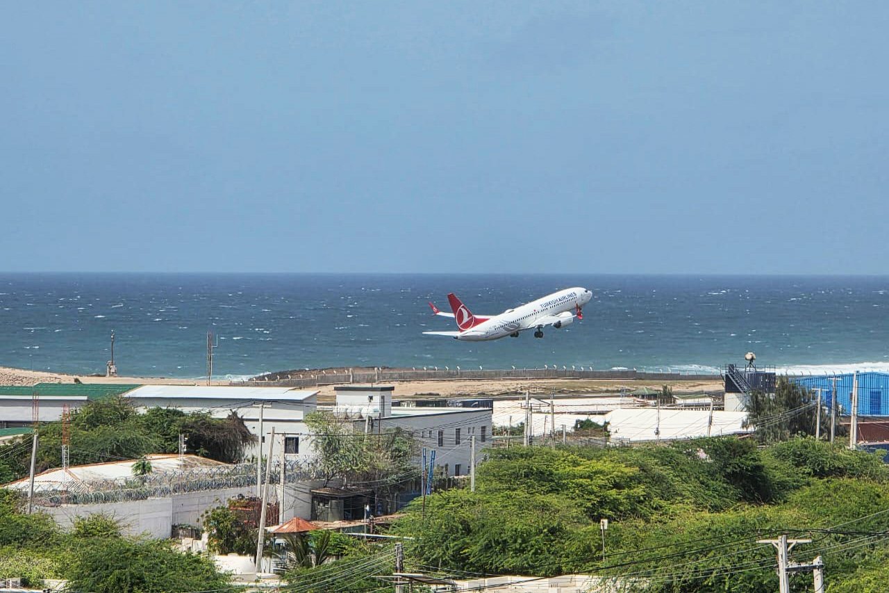 A plane takes off over low buildings and trees along Mogadishu's coastline, with the water on the horizon in the distance beneath a light blue sky.