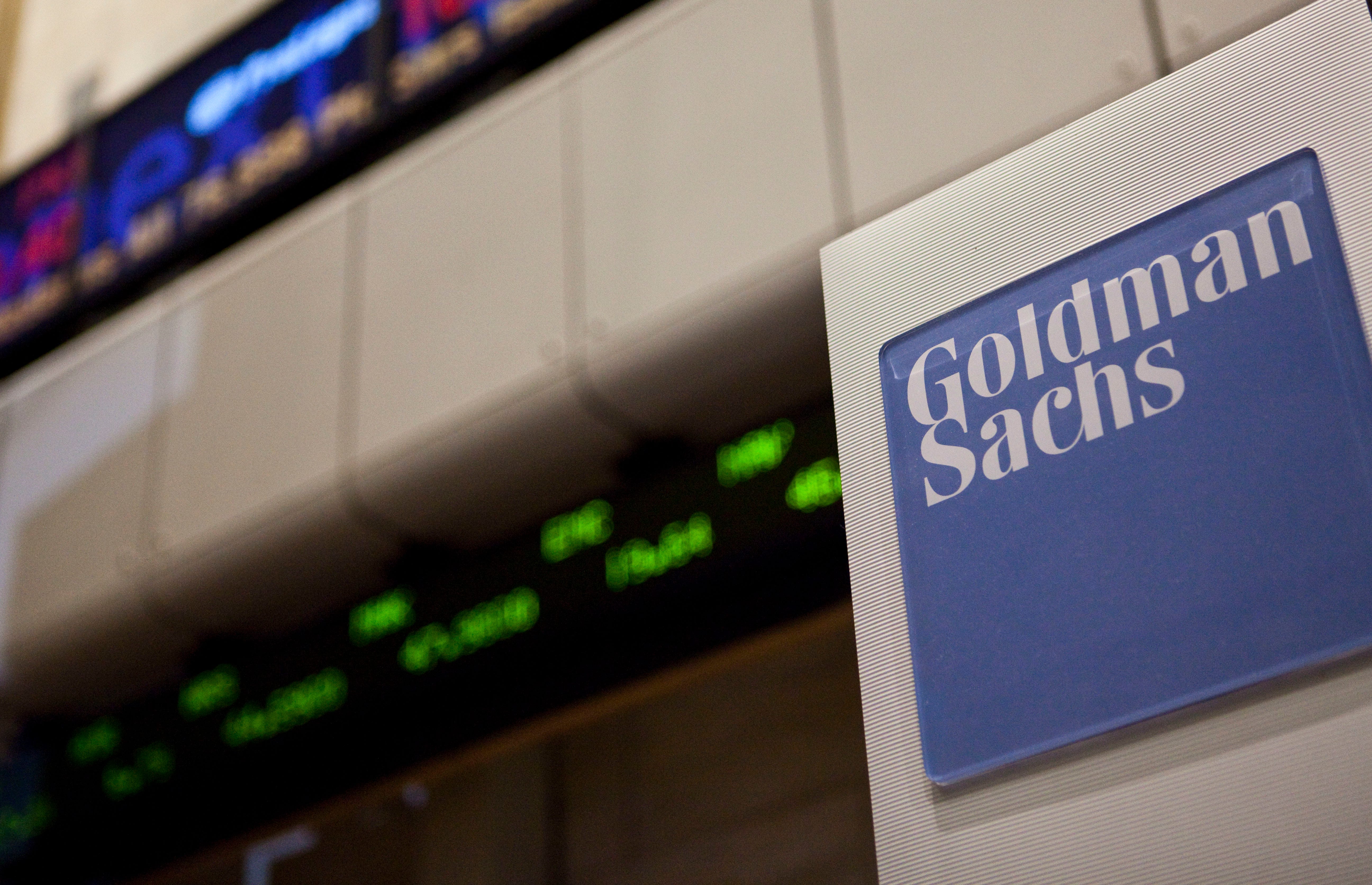 Goldman Sachs logo against backdrop of stocks on screens.