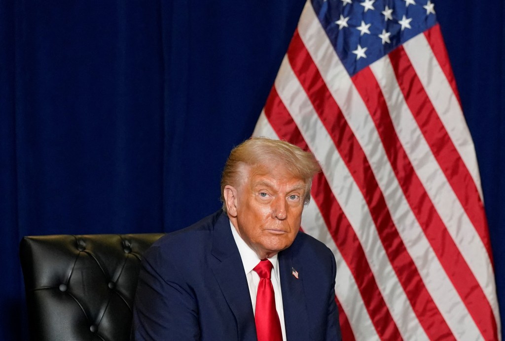 U.S. President Donald Trump in a blue suit and red tie sits in a black leather chair in front of a blue background and a U.S. flag.