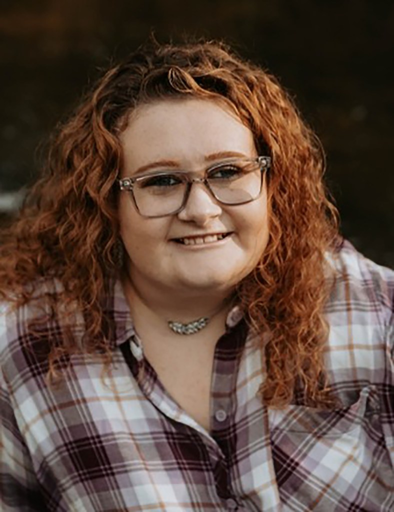Melody Marie Hoffman, a young woman with curly red hair, glasses, and a plaid shirt, smiling at the camera.