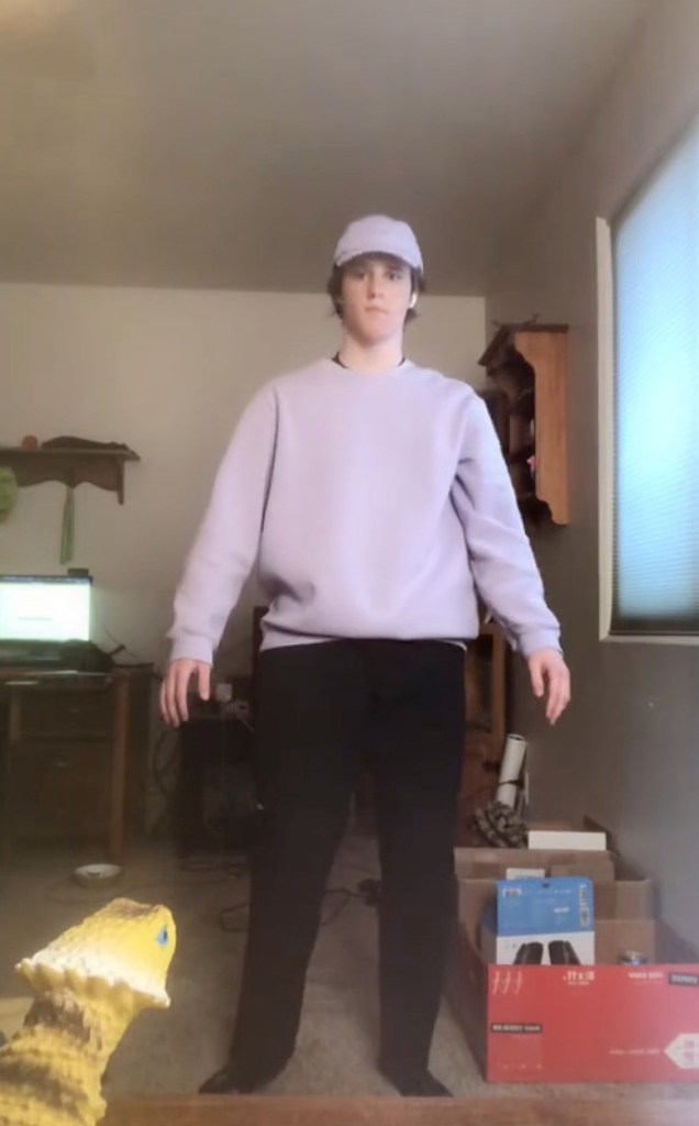 A person in a light purple sweatshirt, black pants, and a light purple hat standing with a toy dinosaur in the foreground.