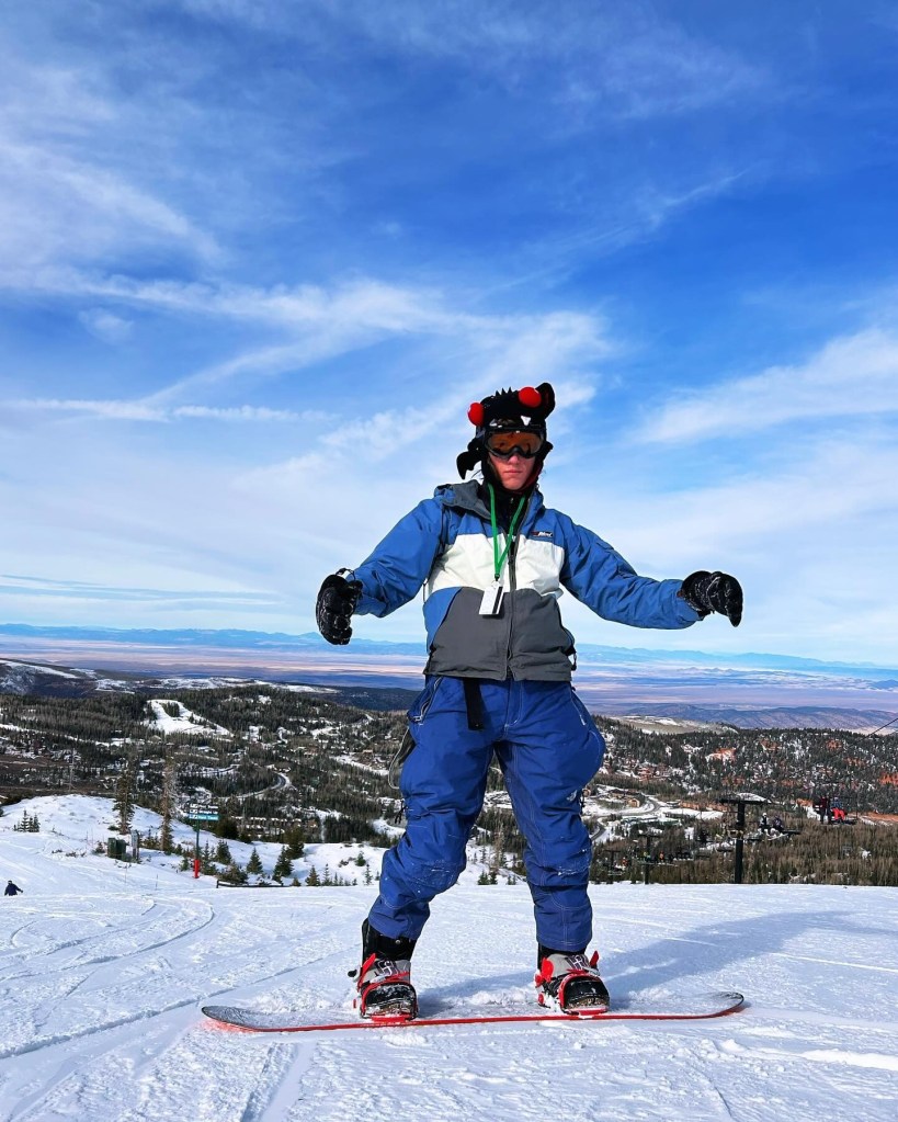 A person wearing a blue and white snow jacket, blue snow pants, and a black helmet with red pom-poms stands on a snowboard at the top of a snowy mountain, overlooking a valley.