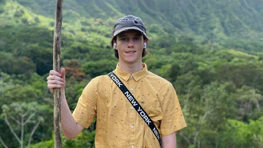 Lance Twiggs, Tyler Robinson's roommate, holds a stick in front of a green, mountainous background.
