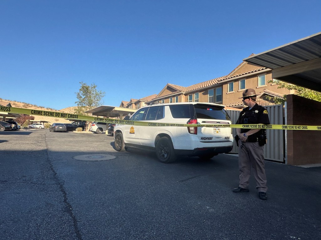 A Utah State Police officer stands guard behind crime scene tape with a police SUV and apartment buildings in the background.