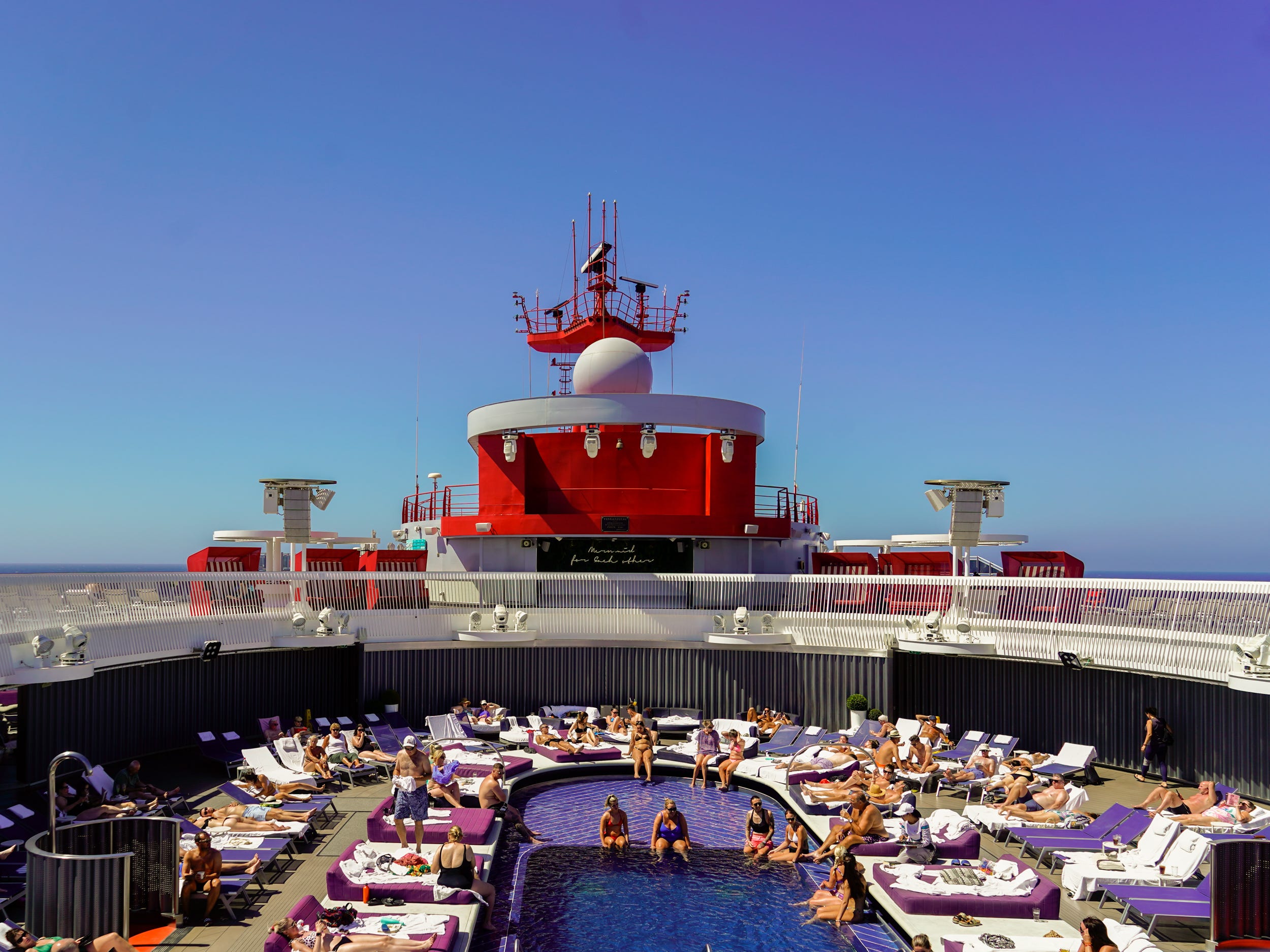 A pool on a cruise ship with people inside it and on pool chairs around it. The sky is clear and blue behind the ship.