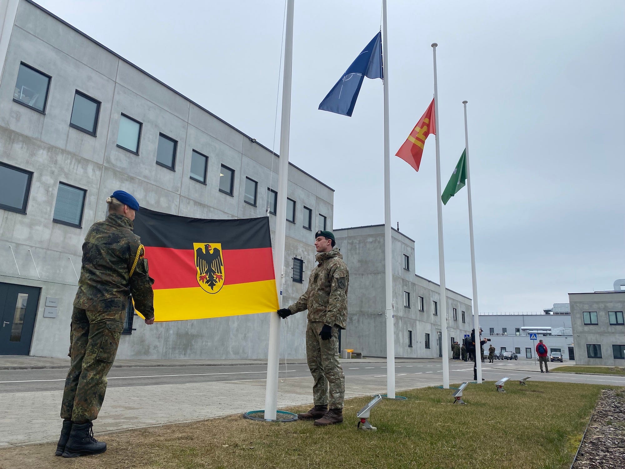 Two soldiers in camouflage gear raise the German flag on a flagpole in front of a gray building.