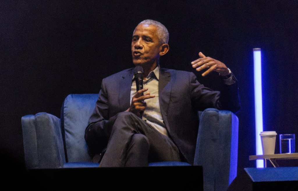 Barack Obama on stage, speaking into a microphone while seated in a blue armchair.