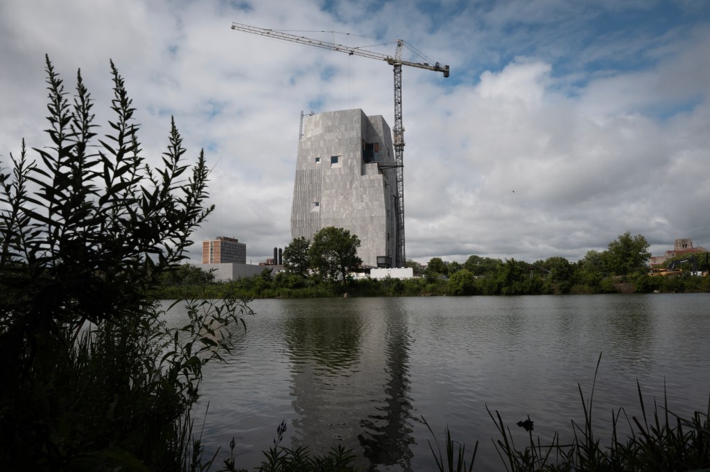 Construction of the Barack Obama Presidential Center in Chicago, viewed across a body of water, with a crane visible next to the unfinished building.
