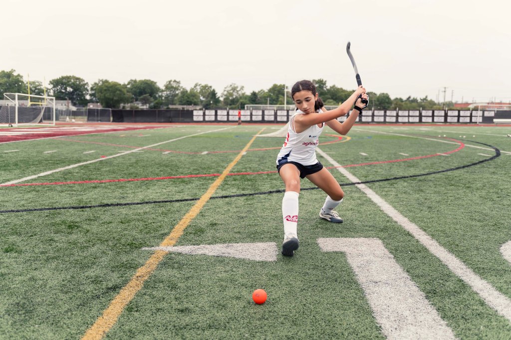 Olivia Goncalves playing field hockey on a field, holding a stick, and positioned to hit the orange ball.