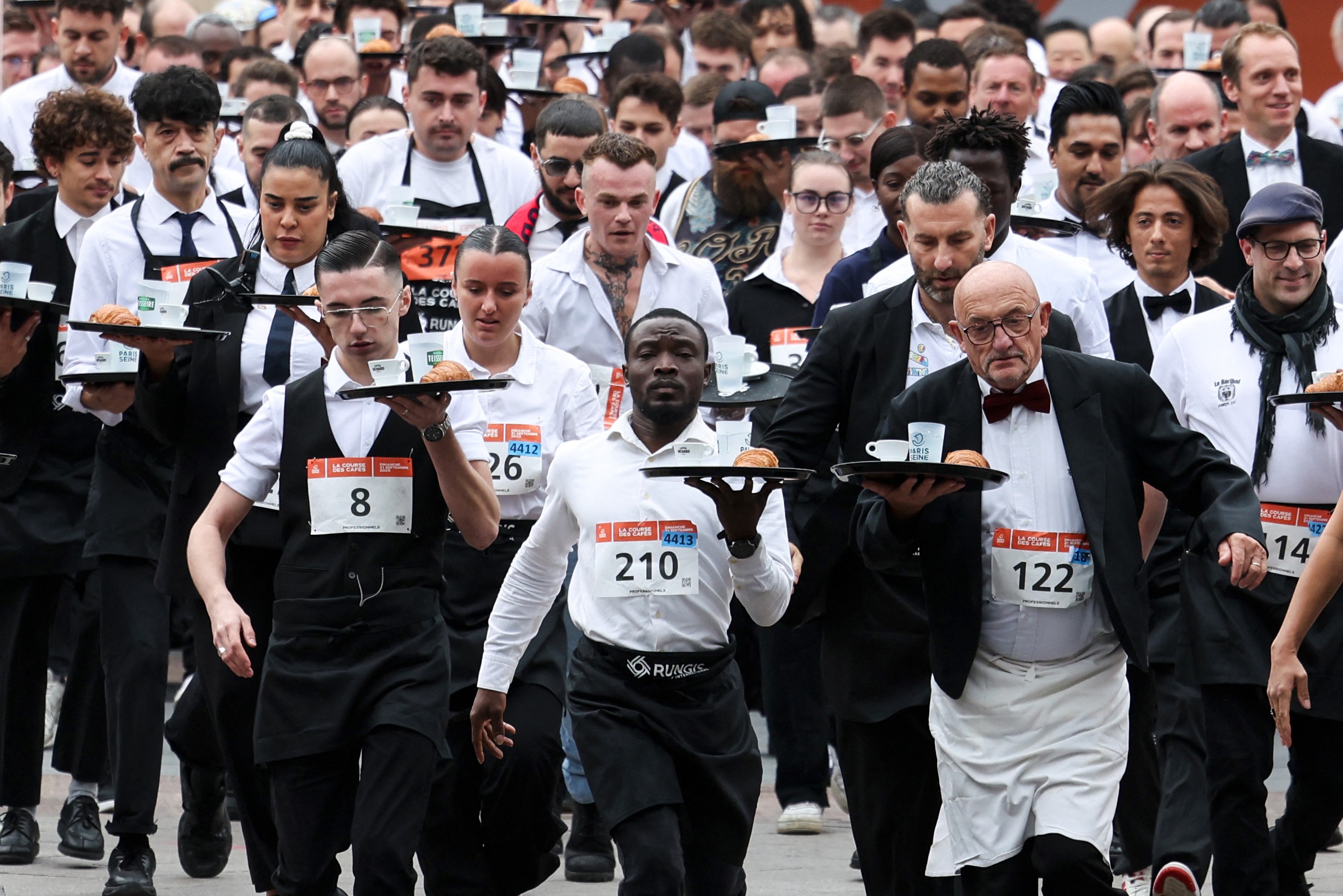 A group of waiters and waitresses, dressed in their work outfits, run a race while carrying a tray.