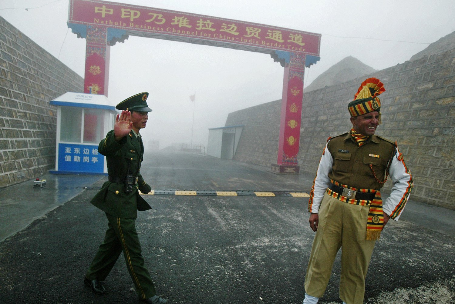 A chinese soldier in uniform holds up one hand as an Indian soldier in uniform laughs in front of a border gate and wall.