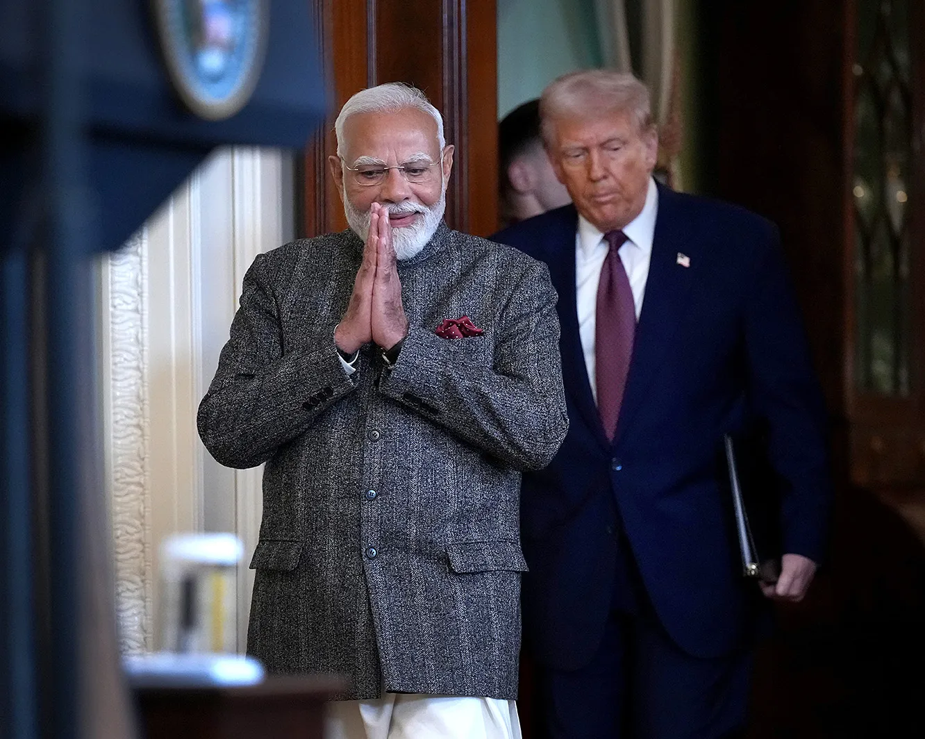 Narenda Modi folds his hands in front of his face as he walks in front of Donald Trump, carrying a folder under one arm, toward a lectern.