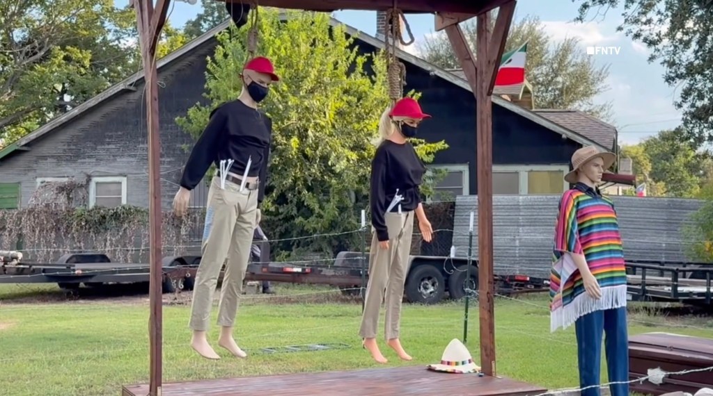 Two mannequins in red hats and black masks hang from a gallows in a Halloween display, with a third mannequin in a poncho standing nearby and a Mexican flag visible in the background.