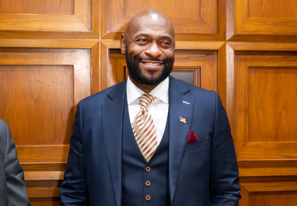 Nathan Wade smiles, wearing a dark blue suit with a striped tie and American flag lapel pin.