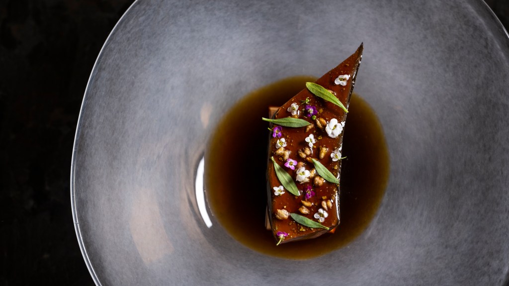 A fine dining dish from Atomix restaurant, featuring a rectangular piece of meat or terrine garnished with edible flowers and herbs, served in a shallow bowl with a rich dark sauce.