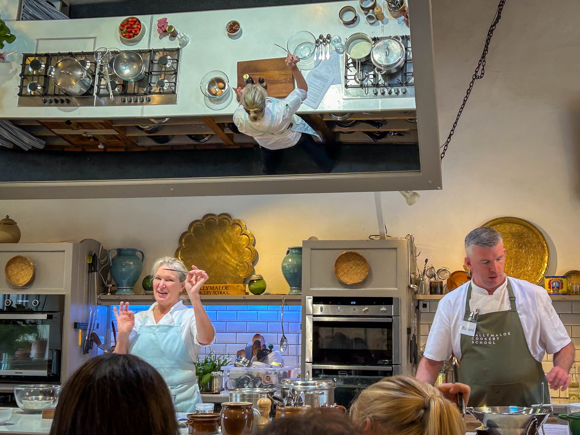 Two chefs giving a cooking demonstration with a large tiled mirror above them.