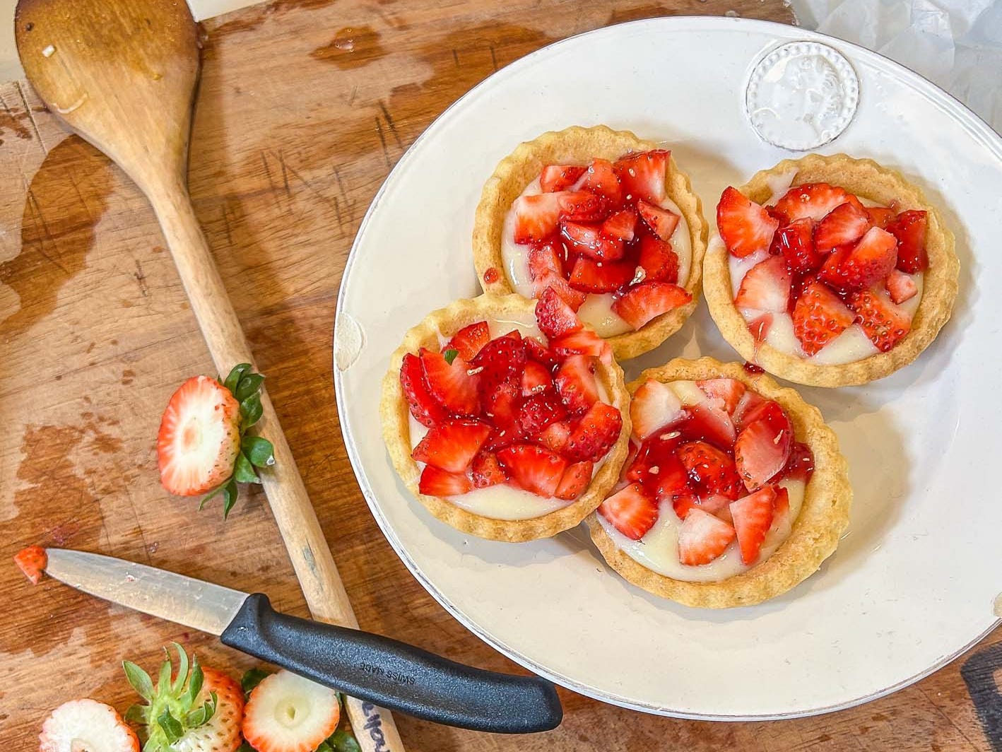 Four strawberry tartlets on a plate next to the heads of chopped strawberries and a wooden spoon.