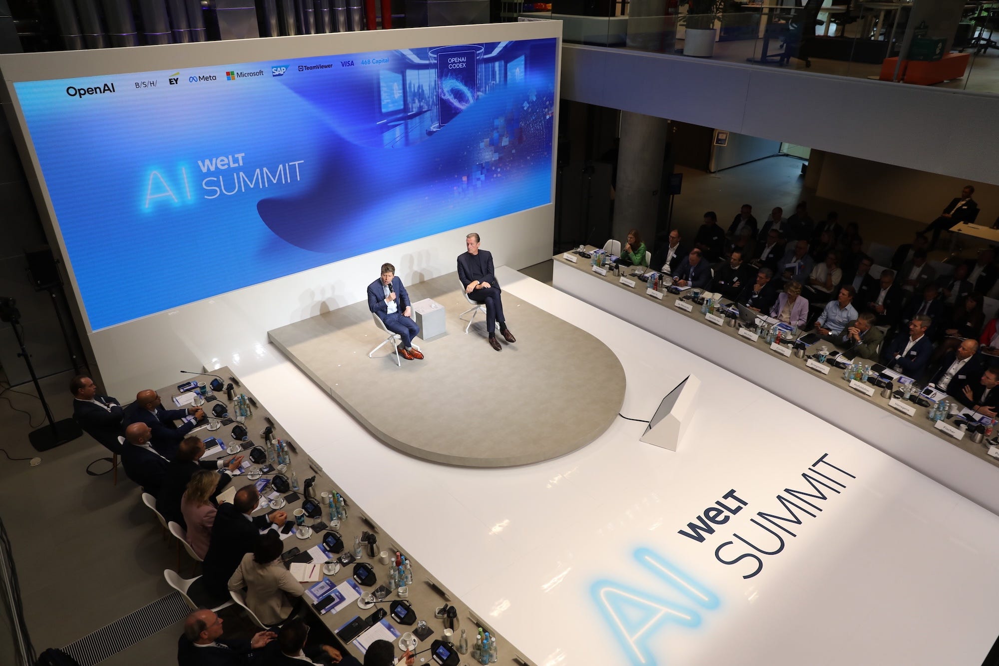 People sit at a U-shaped table curving around a stage.