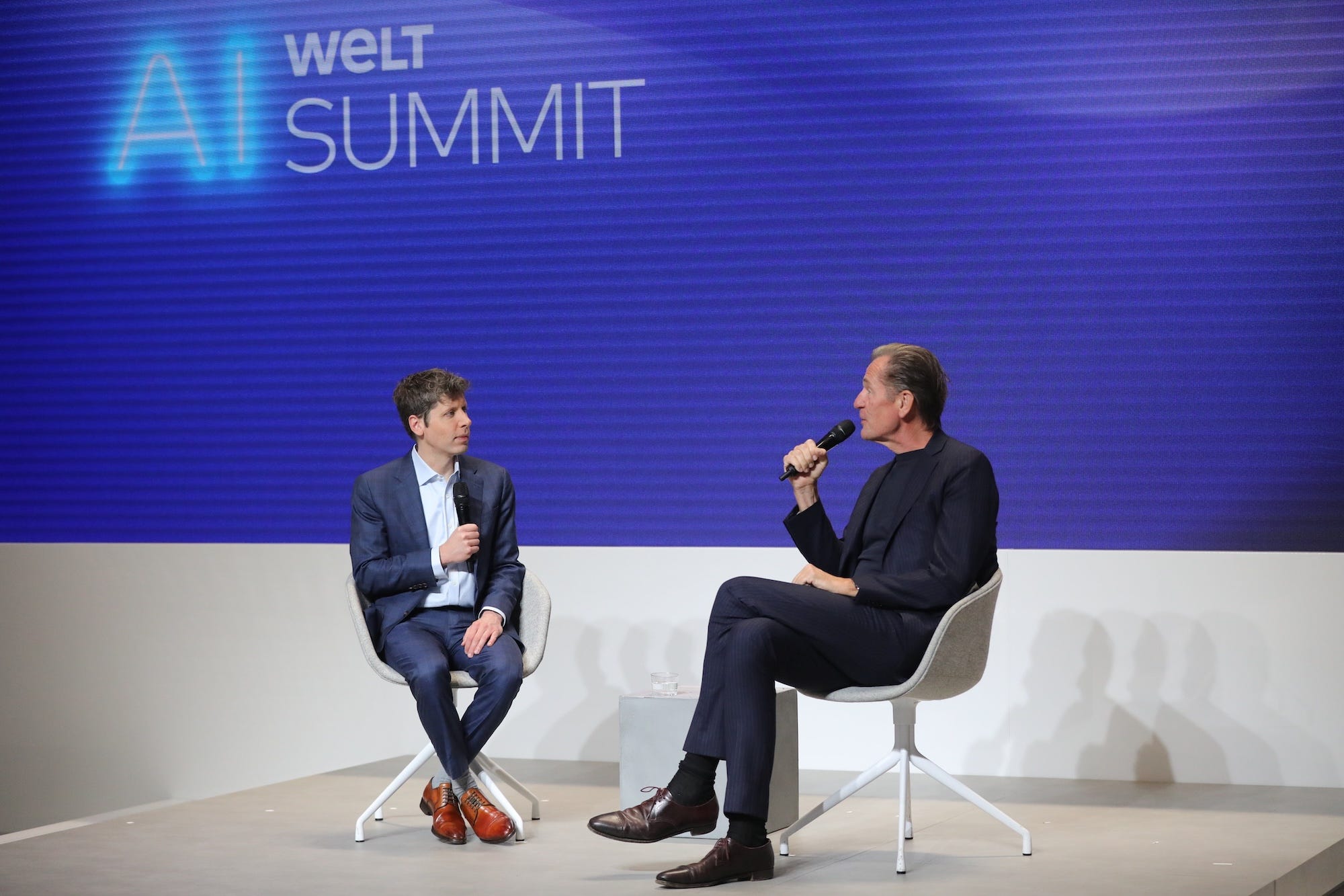 Mathias Döpfner talks into a microphone as Sam Altman listens.