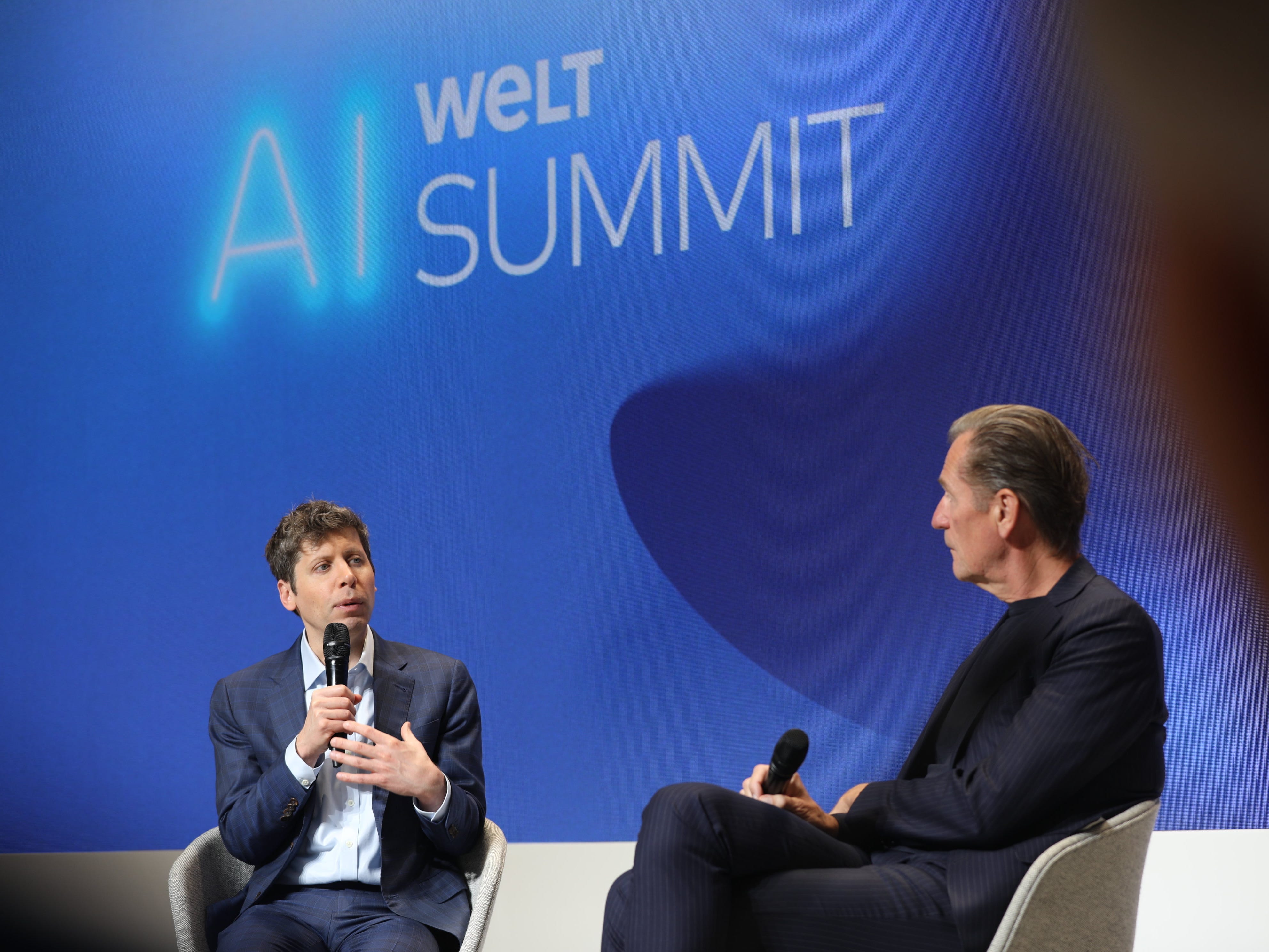 Sam Altman speaks into a microphone and Mathias Döpfner listens intently.