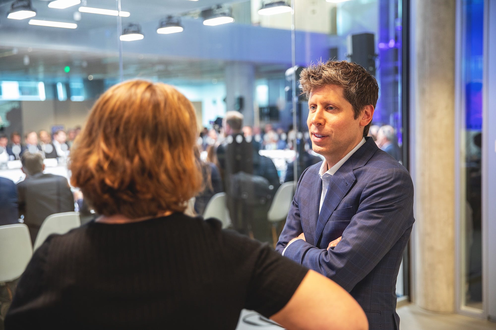 Sam Altman stands with arms crossed, talking to a journalist.