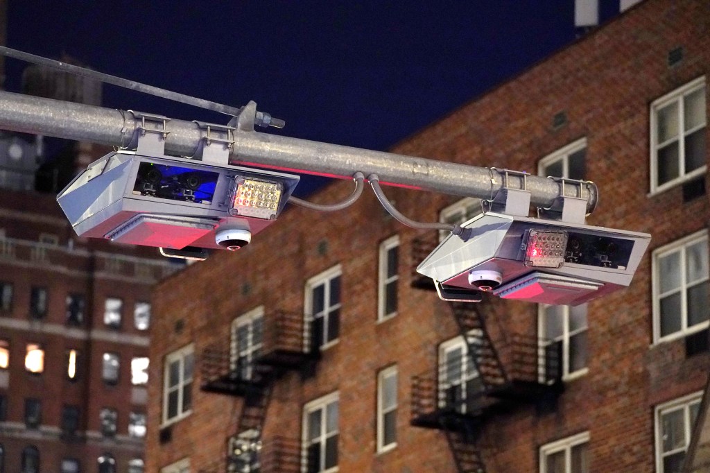 Two congestion pricing cameras and light arrays mounted on a metal gantry, with brick buildings in the background at night.