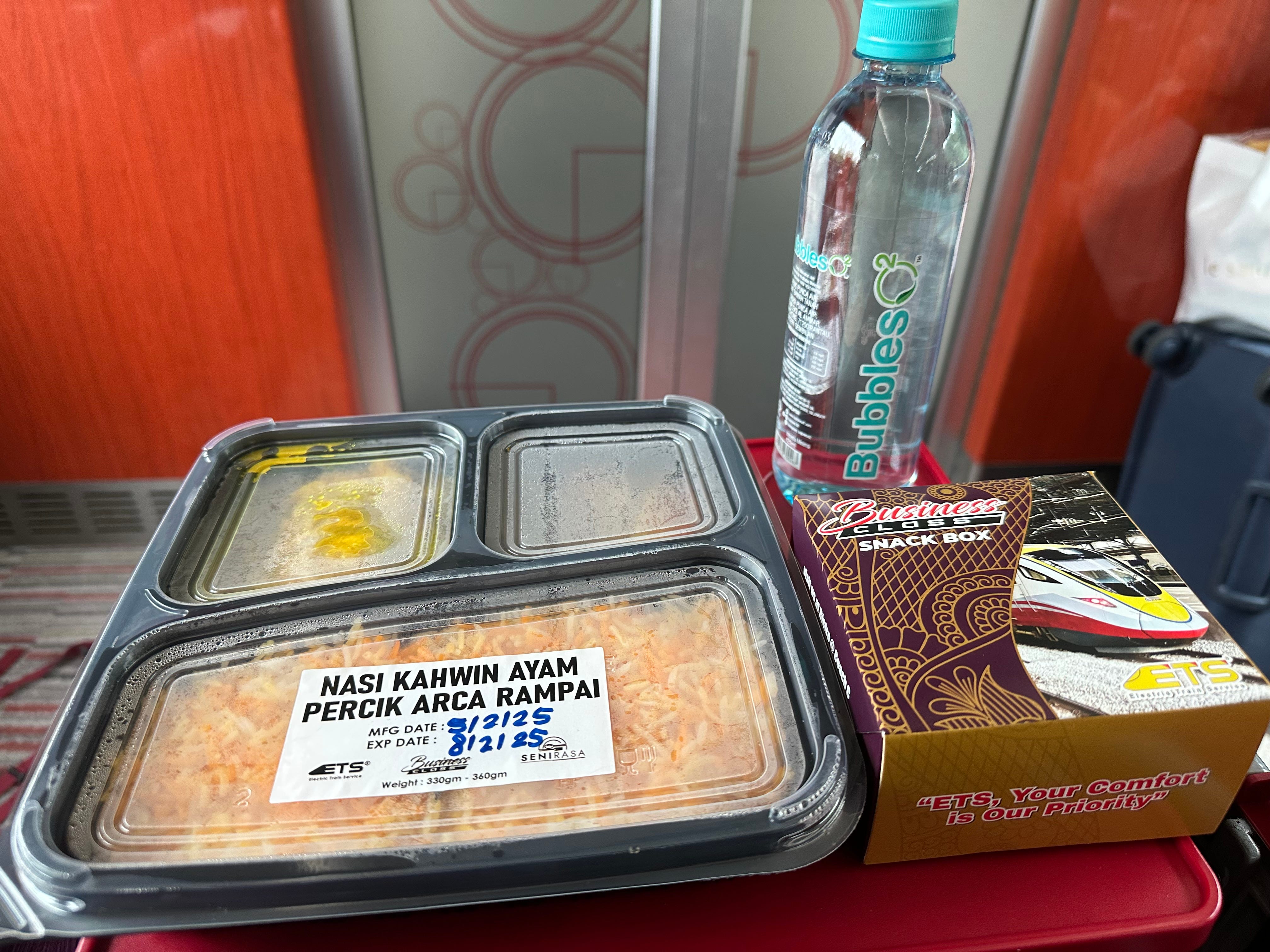 Packed rice meal in plastic container next to snack box, water bottle on tray