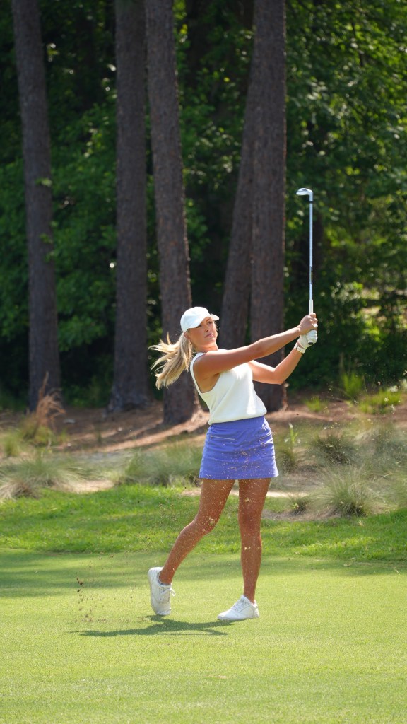 Golf influencer Cailyn Henderson in a white top and purple skirt swings a golf club, sending turf flying.
