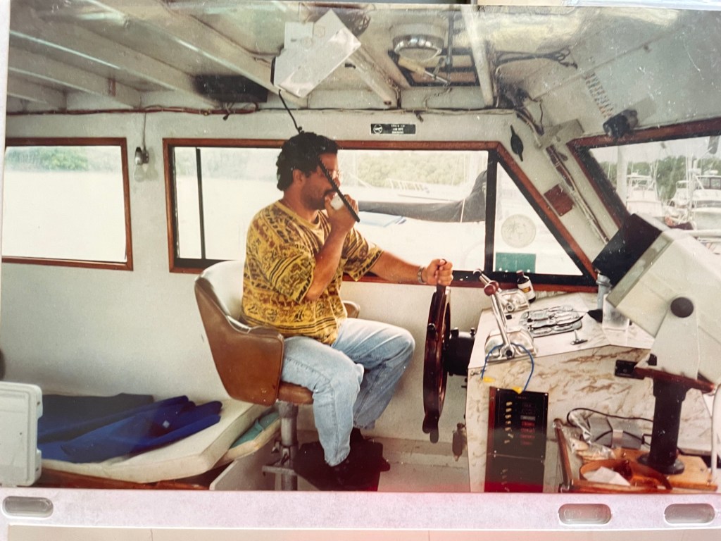 Manny, dressed in a patterned shirt, operates a boat's steering wheel and communication device.