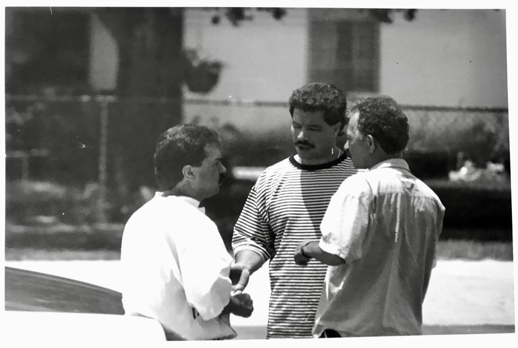 Black and white photo of three men speaking, Martin (center) with a drug trafficker (left) and his sicario (right) from the Medellín Cartel.