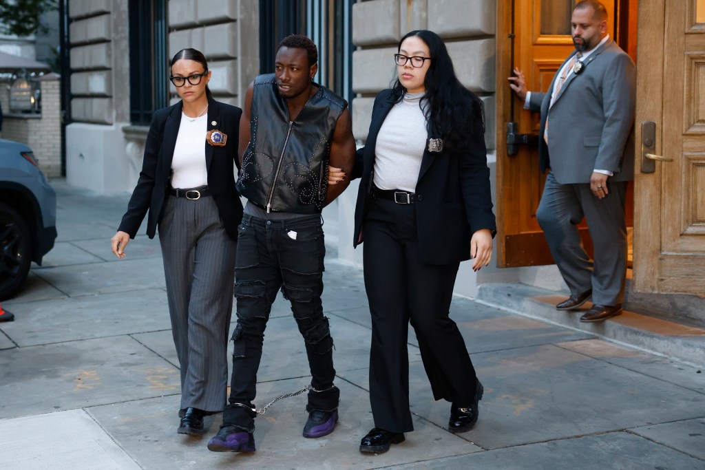 Zion Exum, wearing a black studded vest and black ripped jeans, is perp walked from the Bronx Special Victims Unit, flanked by two officers with a third officer standing in a doorway.