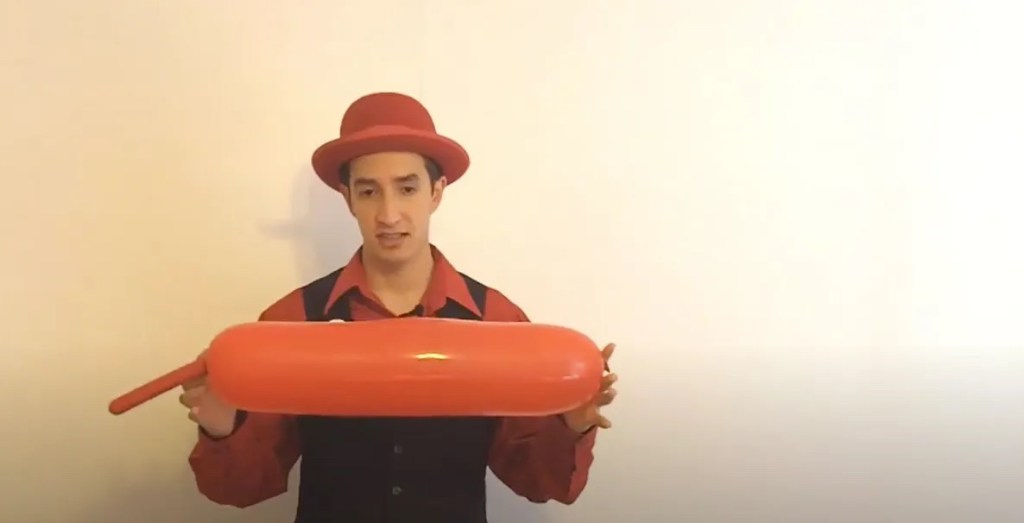 A balloon artist, James Anderson, wearing a red shirt and hat, holds up a long, inflated orange balloon.