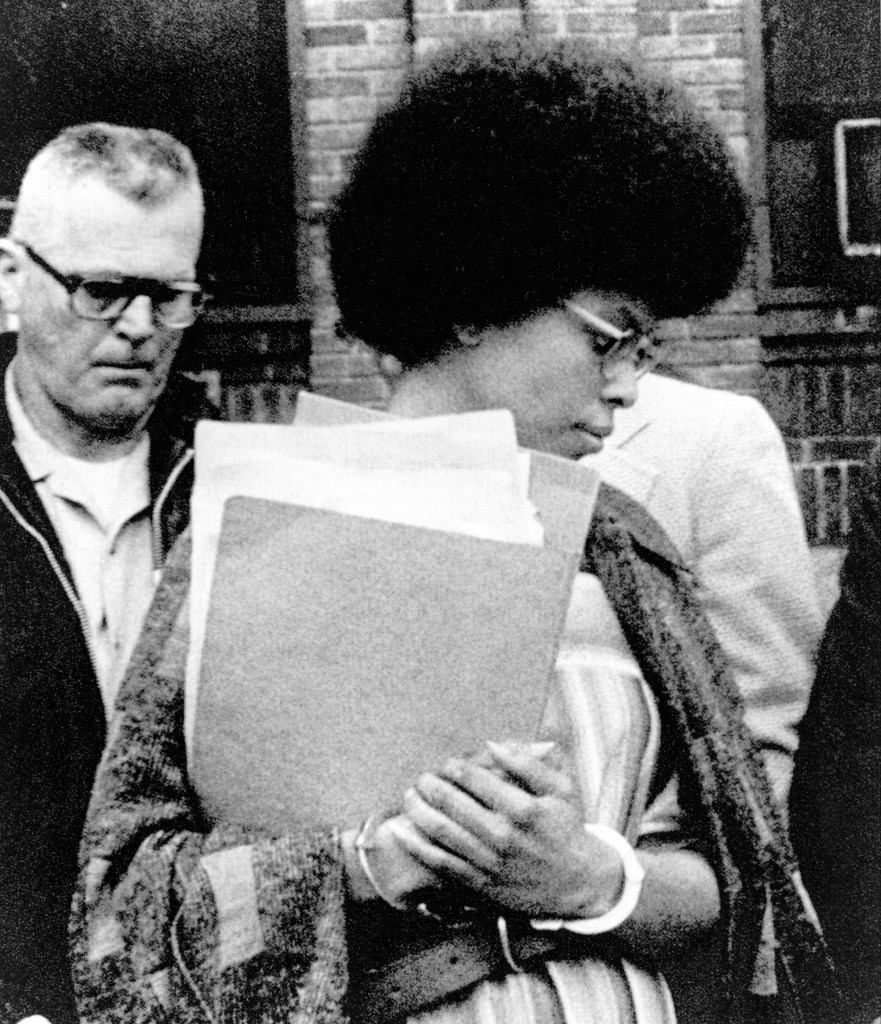 Joanne Chesimard, also known as Assata Shakur, wearing handcuffs and holding files, walks past a man in glasses.