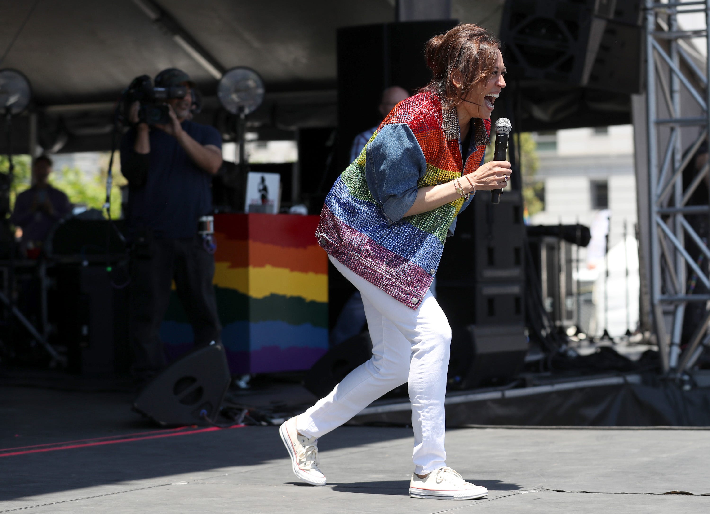 Kamala Harris onstage, laughing and holding a microphone. She wears a rainbow jacket, white trousers, and white sneakers.