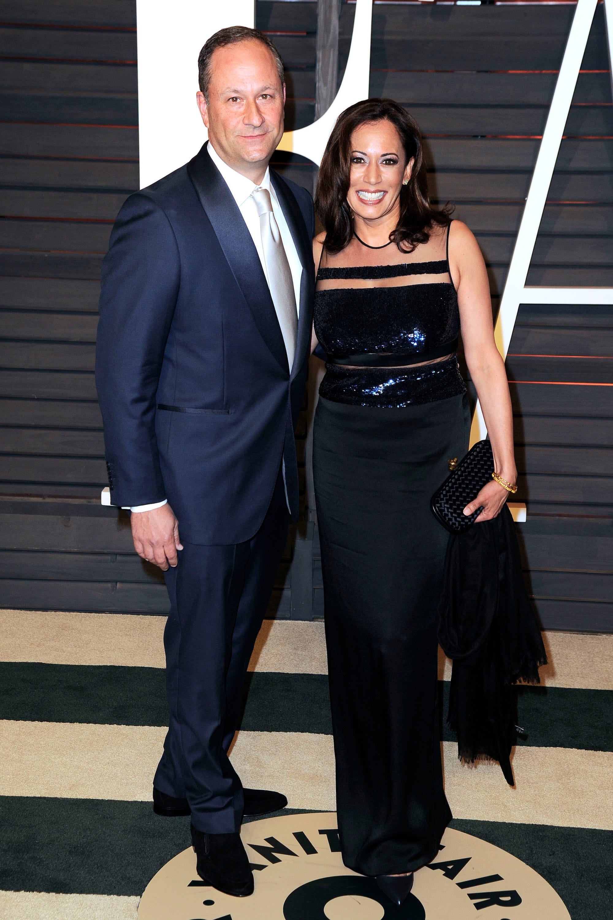 Doug Emhoff and Kamala Harris posing together on the Vanity Fair carpet. He wears a navy suit and silver tie, while she wears a black floor-length dress.