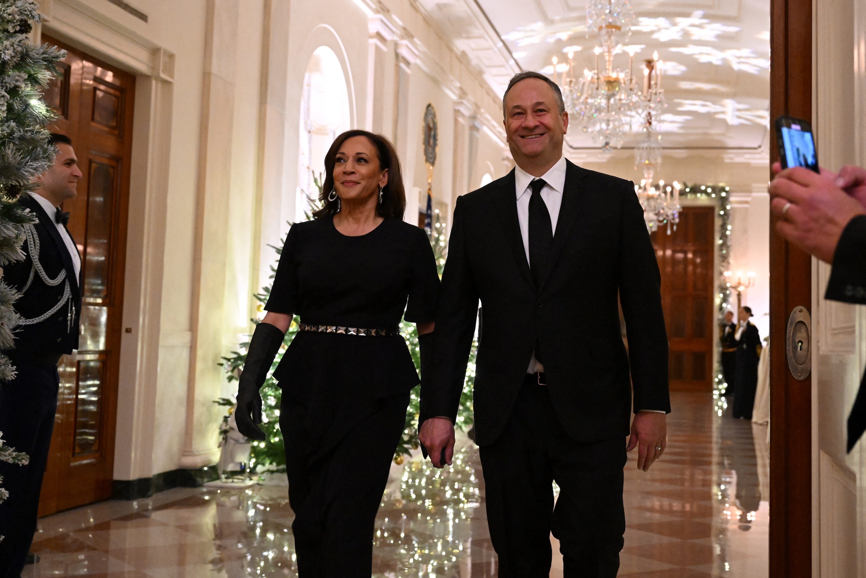 Kamala Harris and Doug Emhoff holding hands and walking together through a grand hall with Christmas decorations. She wears a black outfit and black gloves, and he wears a black suit and tie.