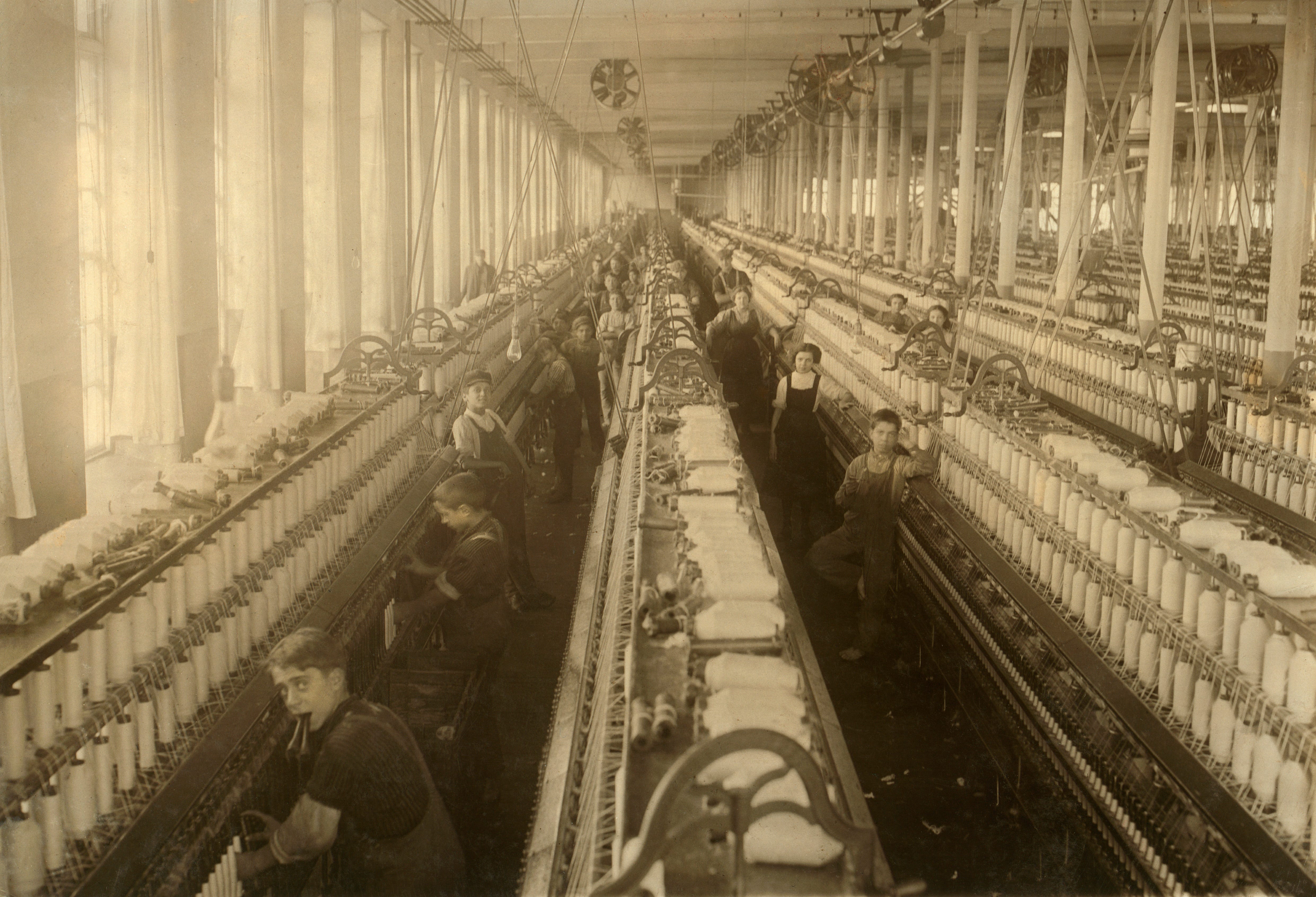 Women and children stand at large spinning machines in a factory