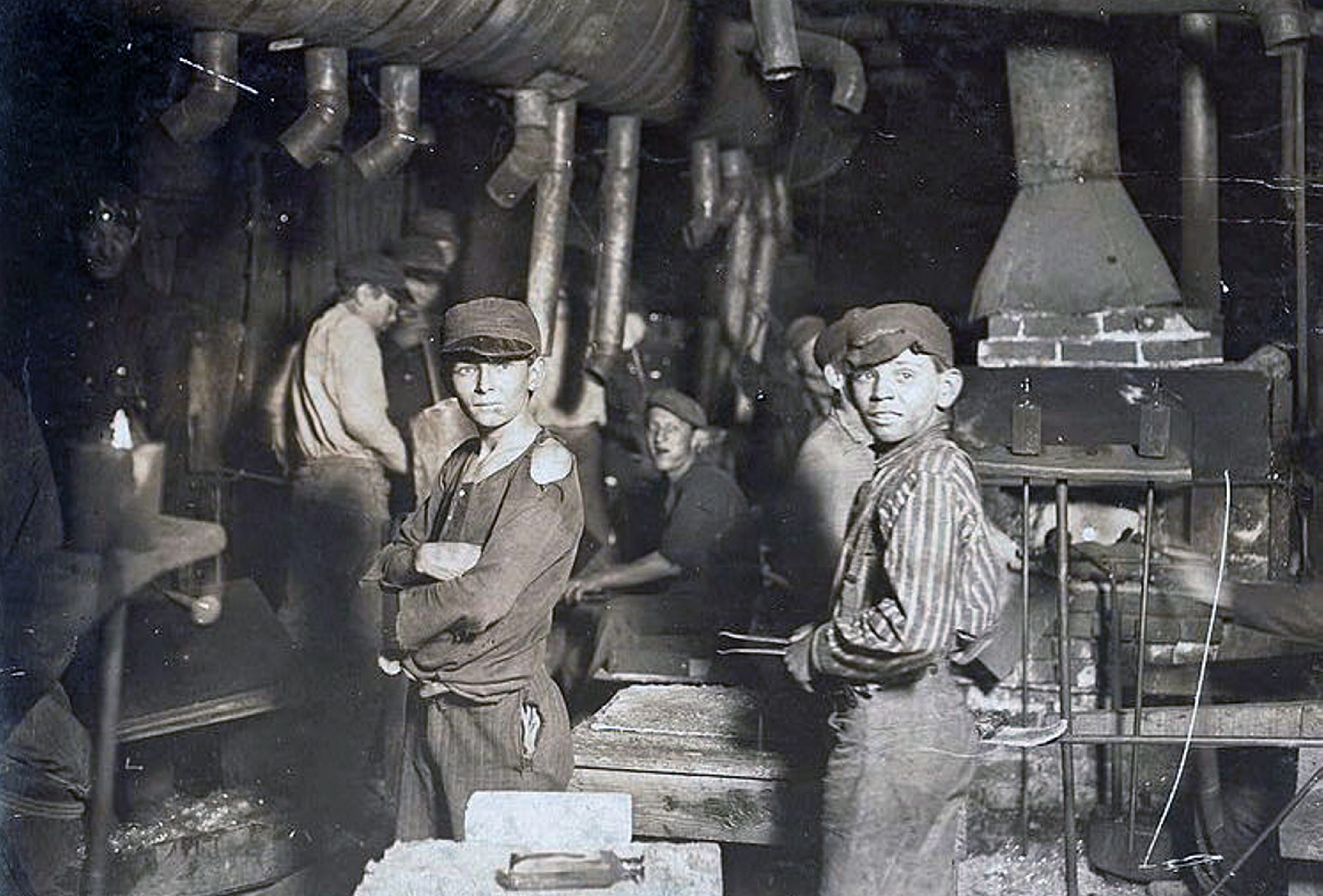 Two boys in hats with dirty faces at a glassworks factory with other workers in the background