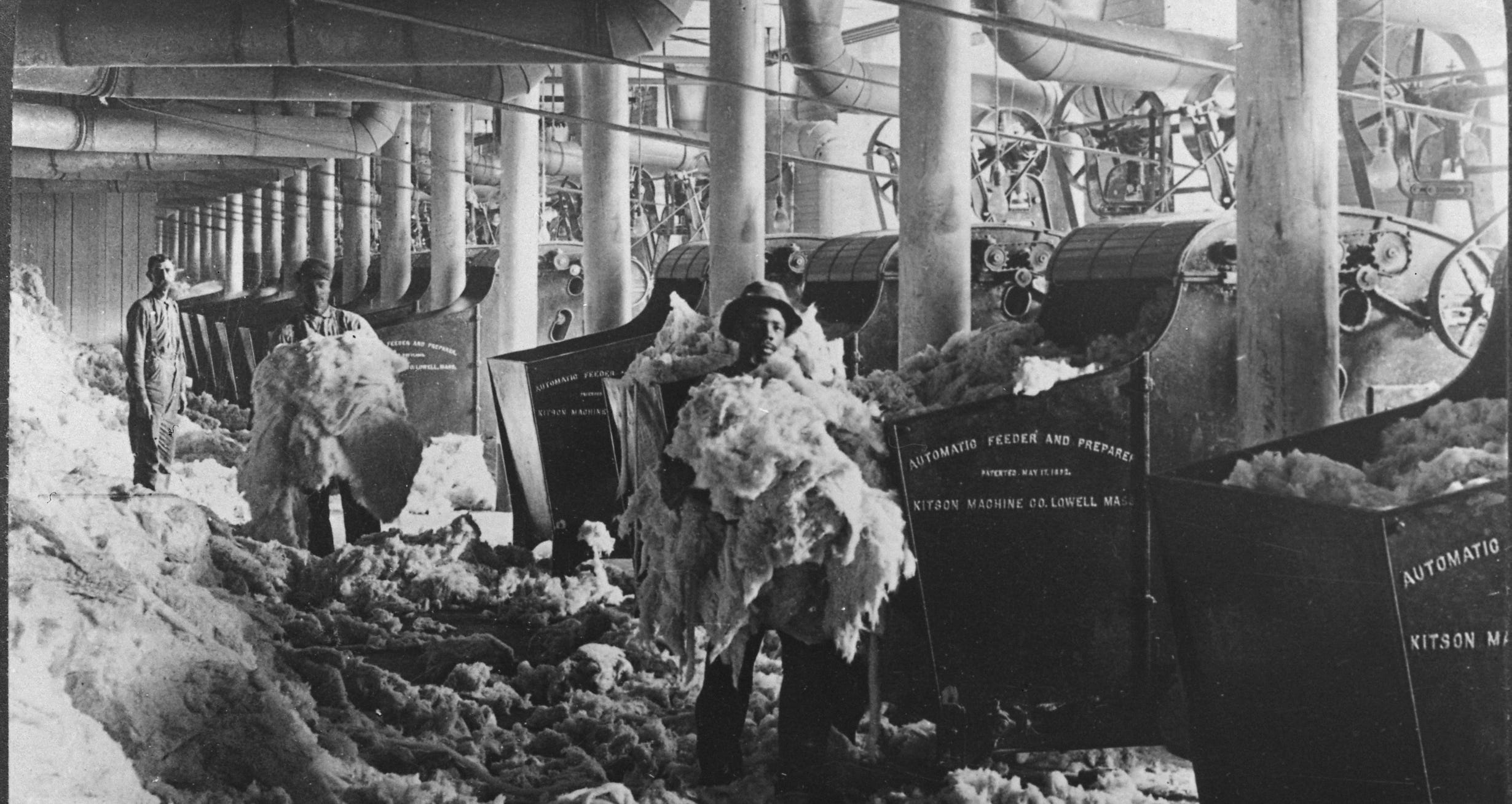 Men hold armfuls of cotton near large equipment