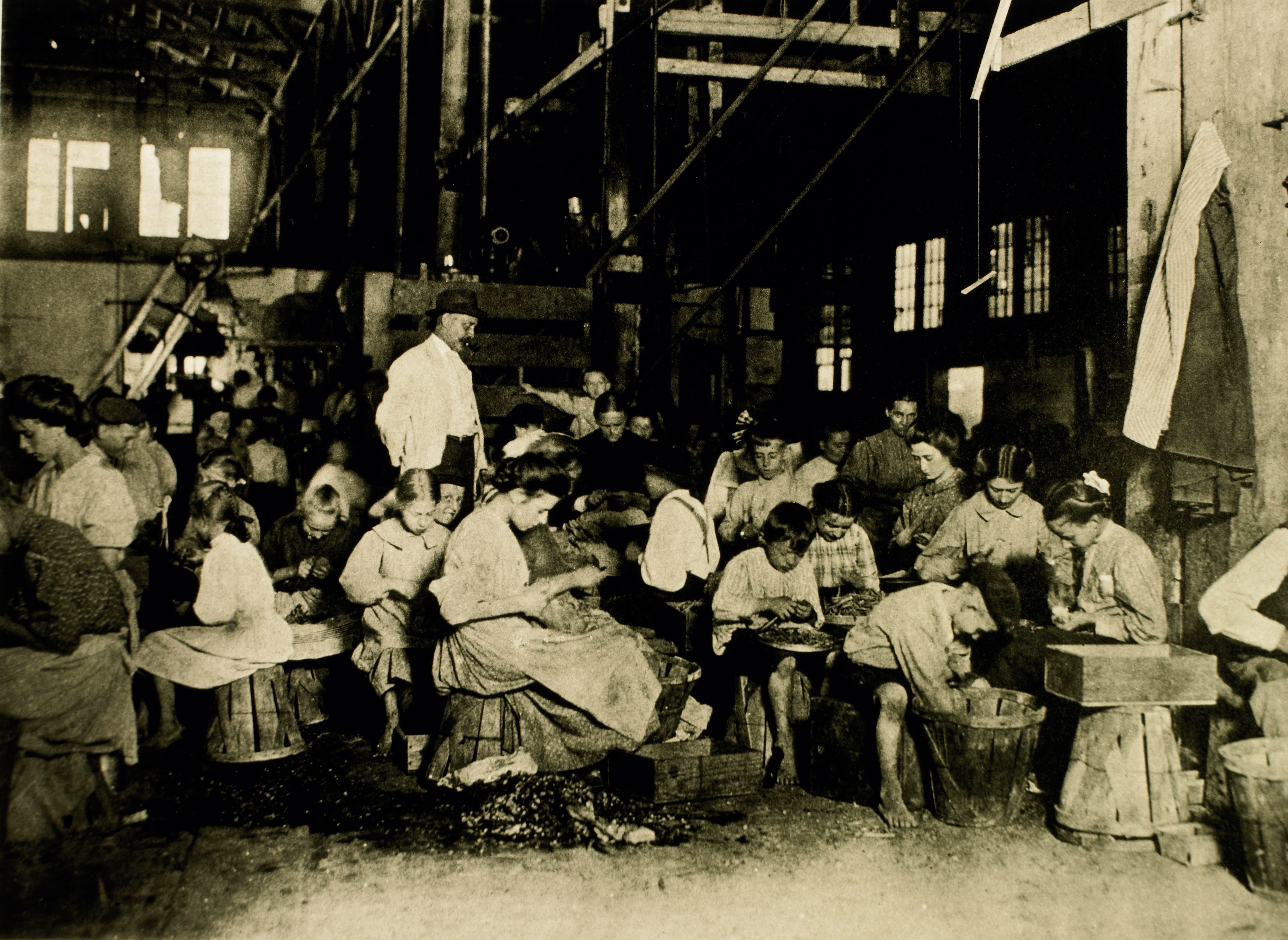 A crowd of women and children sit working in a cannery as a man watches