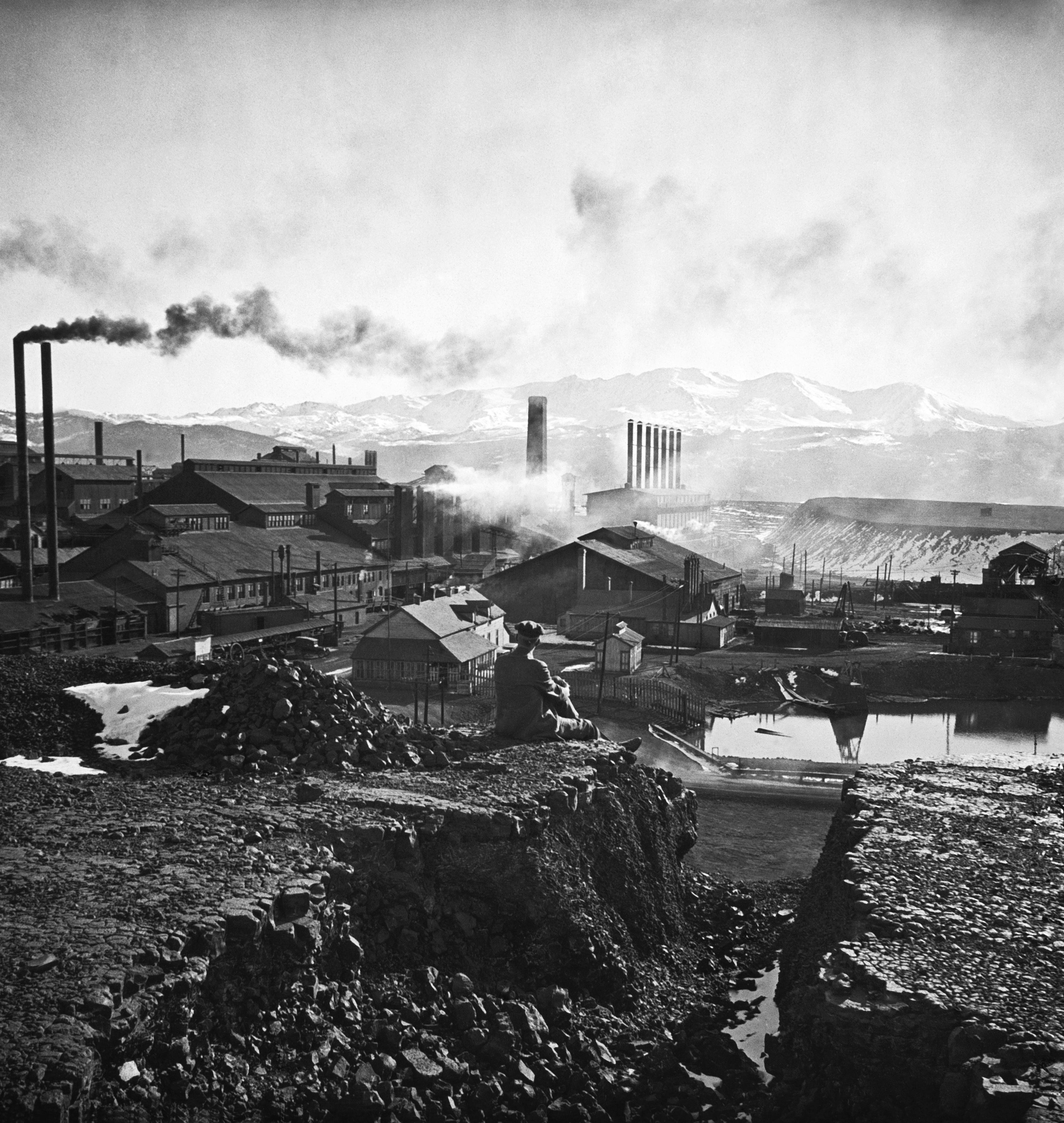 A man sits looking at a lead mill, which is belching smoke