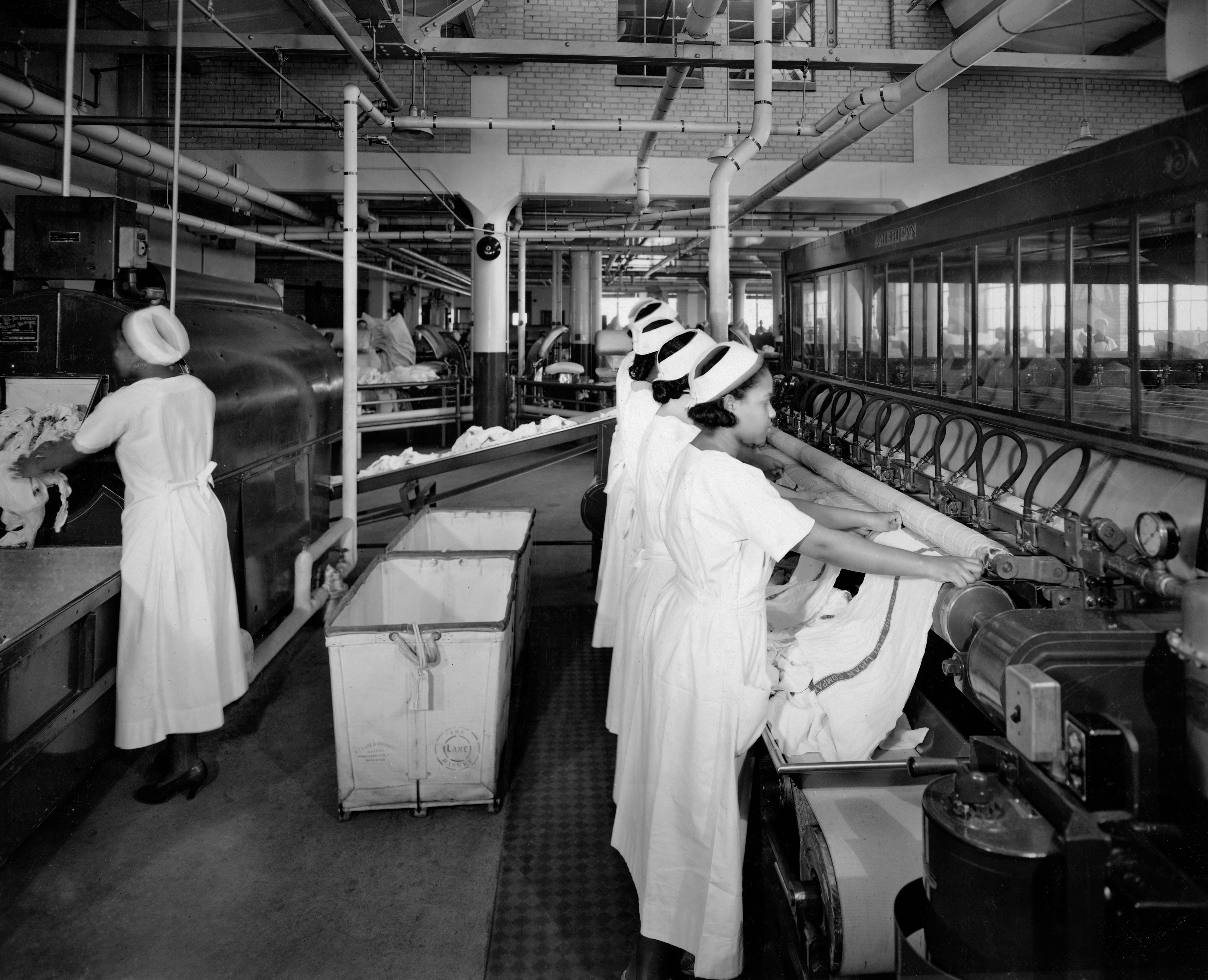 Women in white dresses and caps feed laundry into large machinery