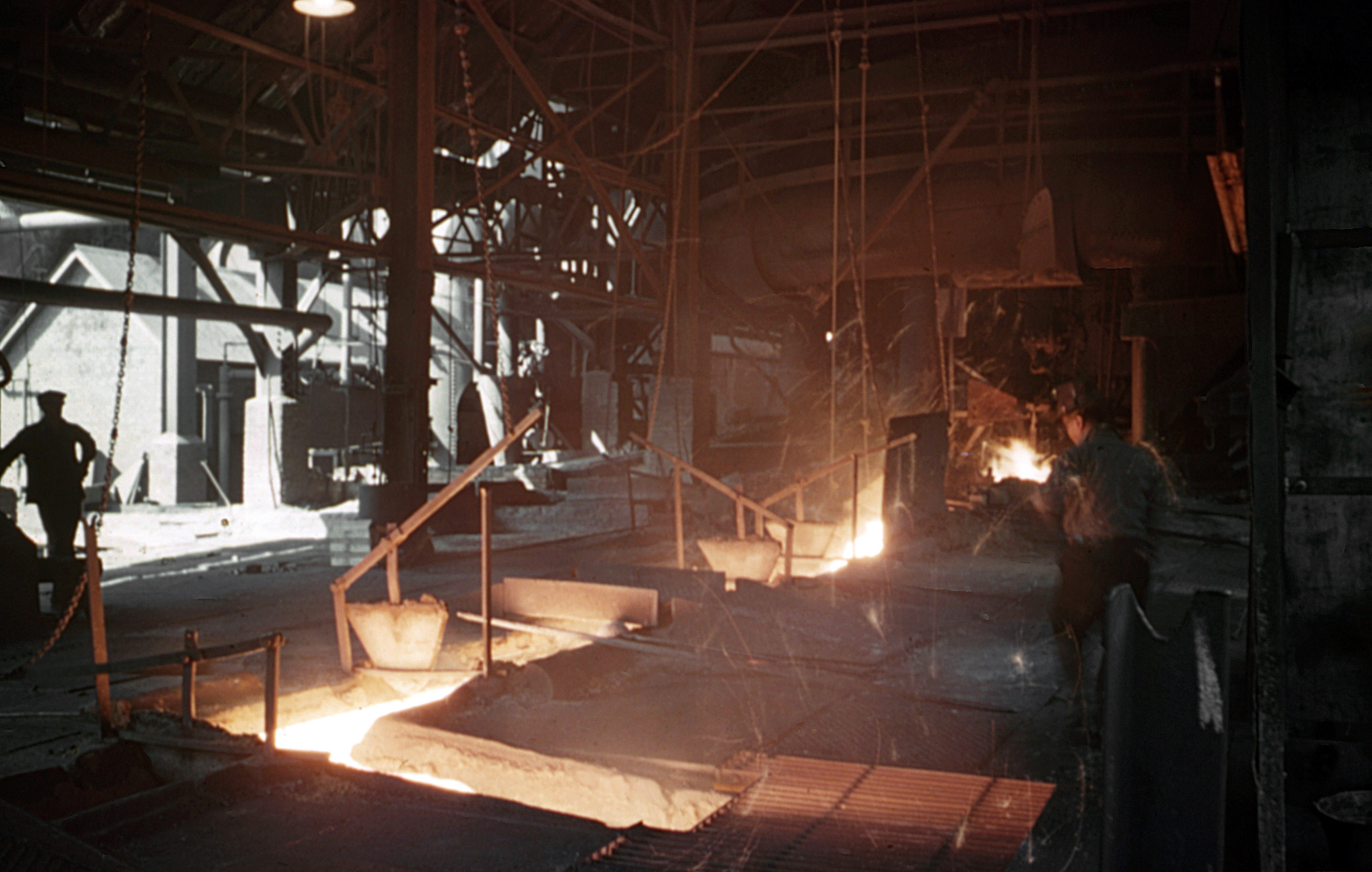 Men work near glowing pits in a steel mill with machinery around