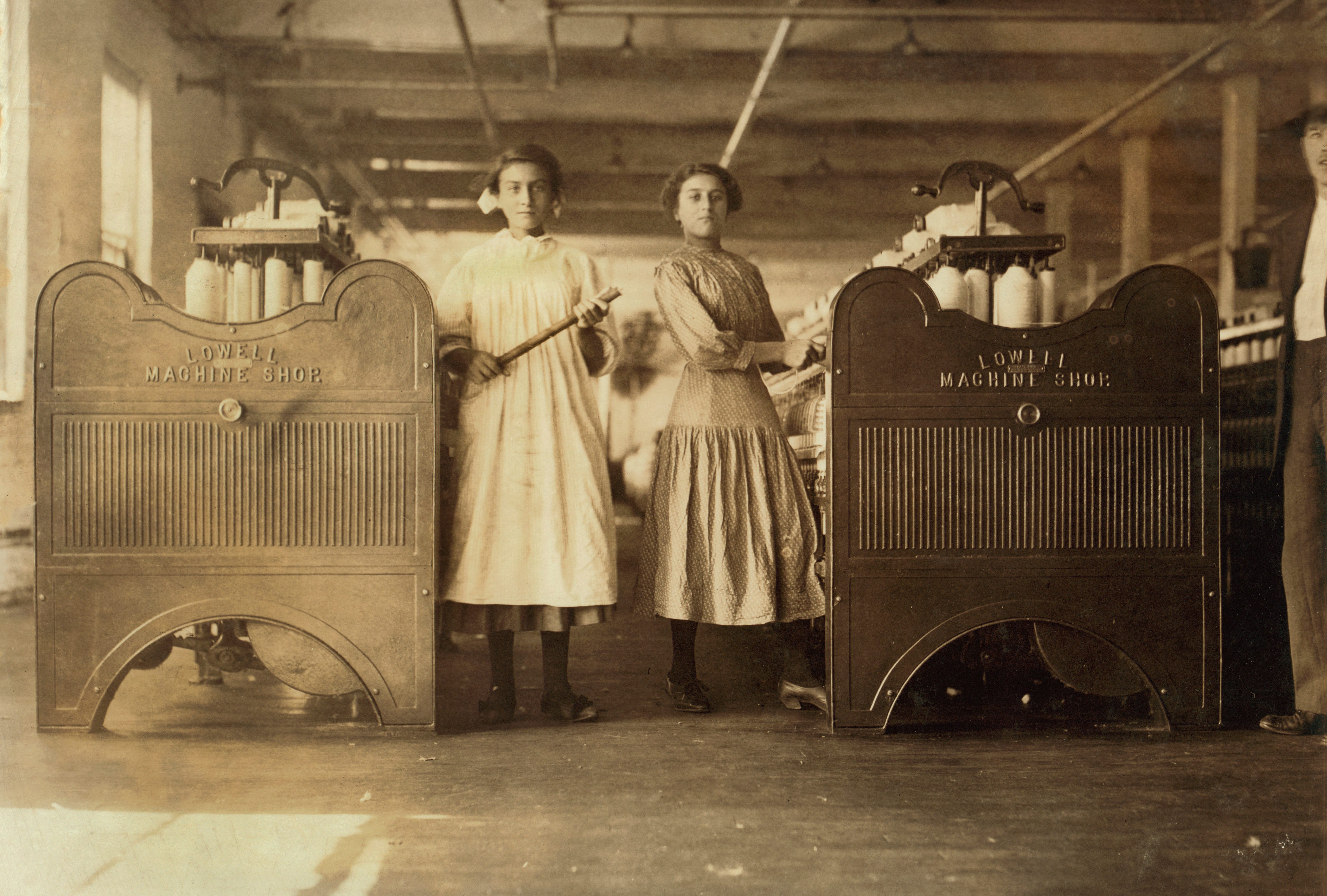 Two women in dresses stand next to factory equipment, one holding a stick
